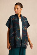 A woman wearing the Abstract Floral Wool Gilet— a dark, open-front cardigan with short sleeves, a shawl collar, and a colorful abstract pattern— layered over a white top and green textured skirt.
