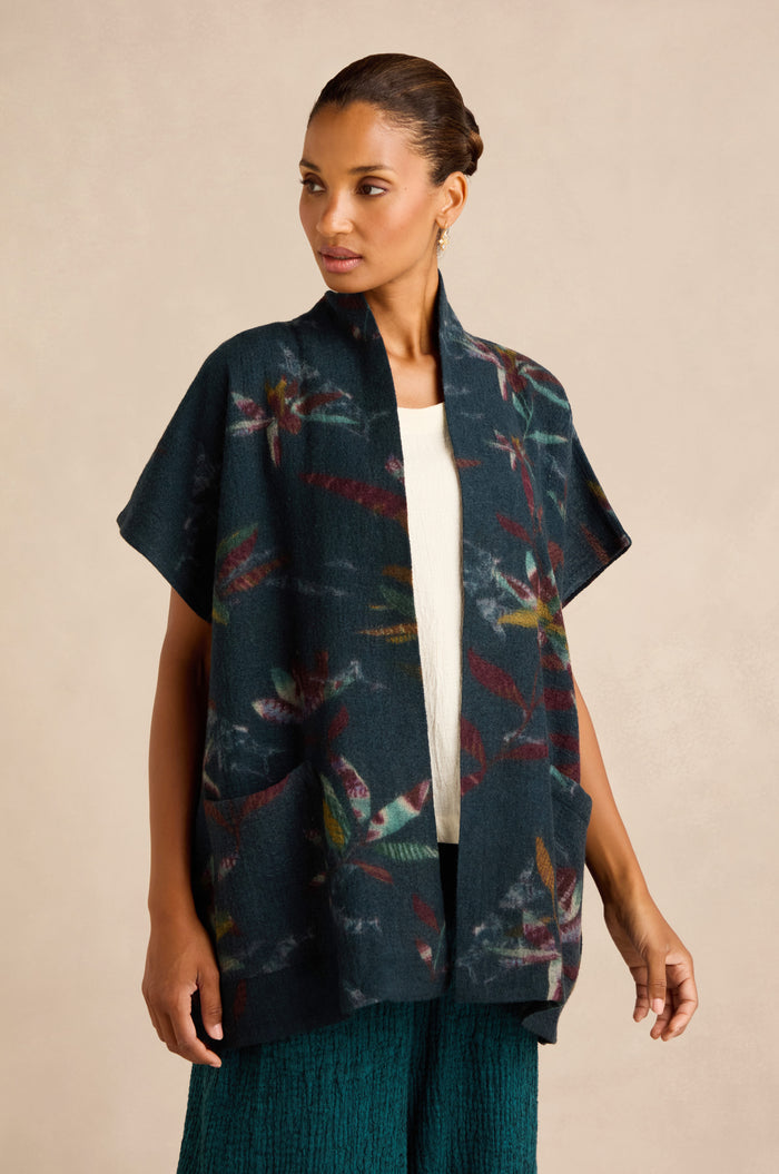 A woman wearing the Abstract Floral Wool Gilet— a dark, open-front cardigan with short sleeves, a shawl collar, and a colorful abstract pattern— layered over a white top and green textured skirt.