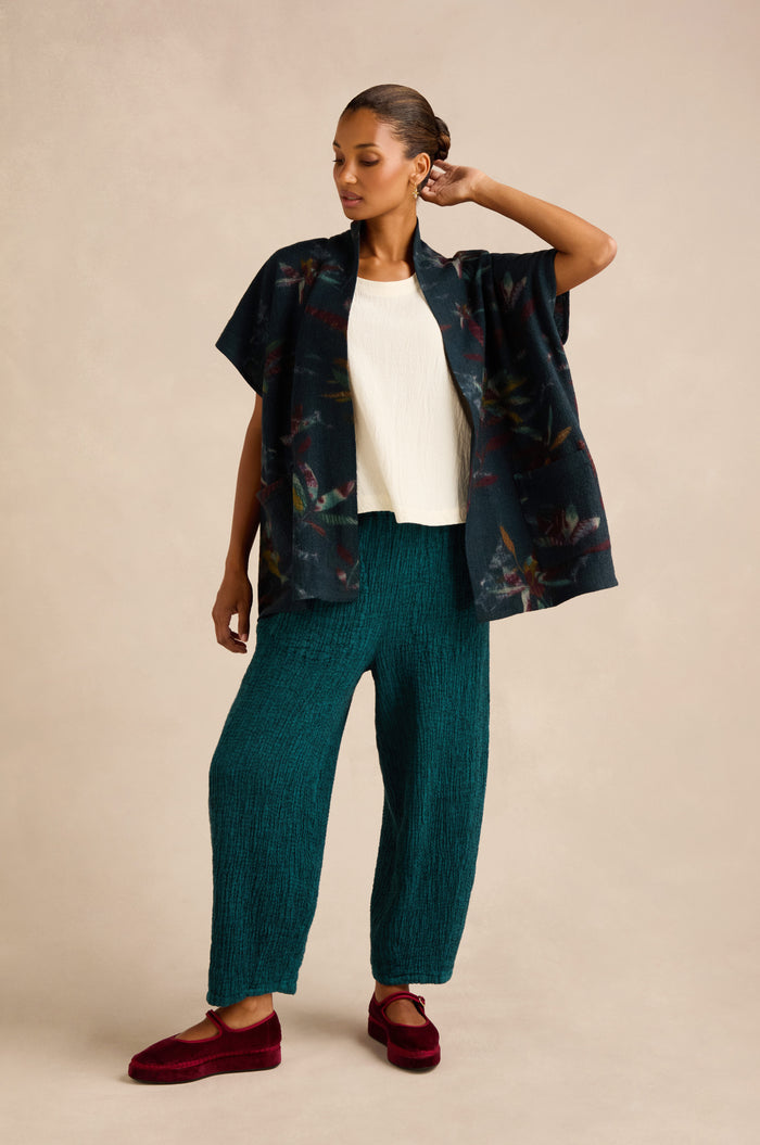 A woman stands against a plain beige background wearing a white top, a patterned dark short-sleeve shirt, textured teal pants, and the Abstract Floral Wool Gilet featuring a shawl collar in brushed wool, paired with red shoes.
