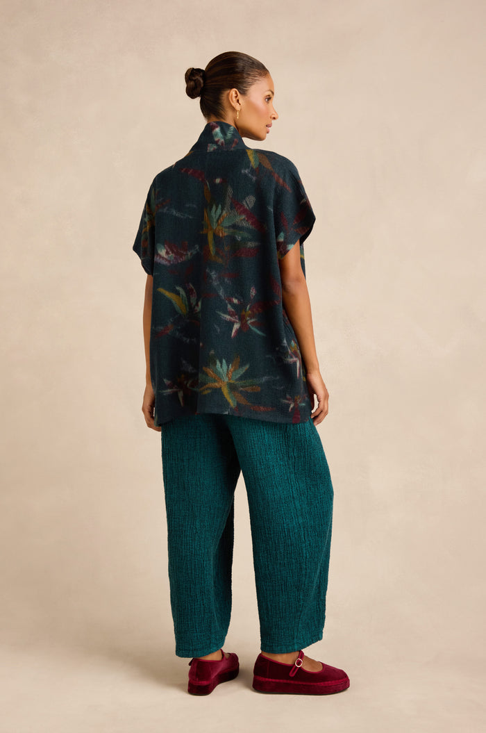 A woman stands facing away, wearing the Abstract Floral Wool Gilet—a short-sleeve, dark patterned top with a shawl collar—paired with teal textured pants and red shoes, against a neutral background.