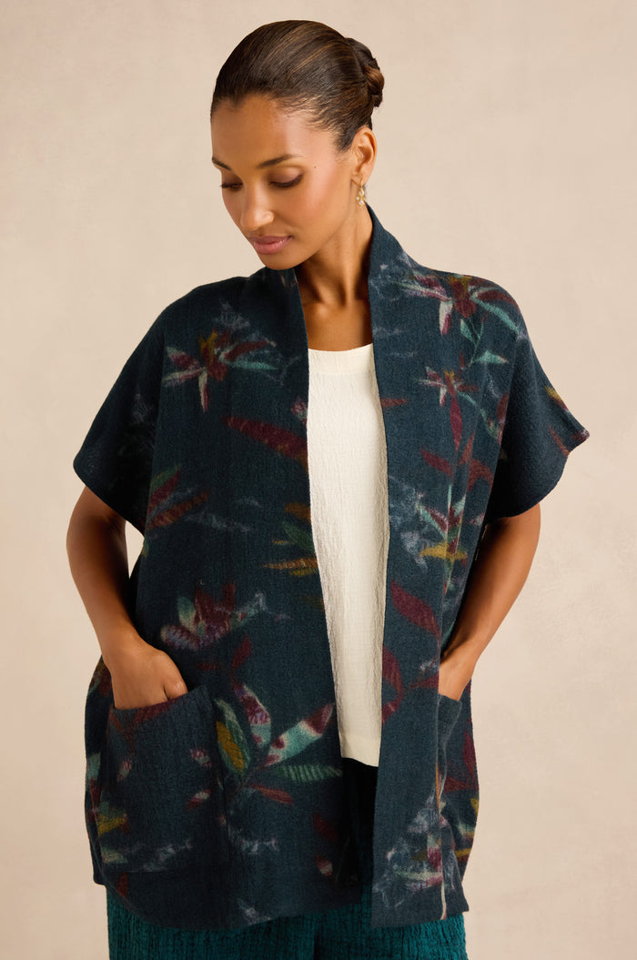 A woman wearing a dark, short-sleeved Abstract Floral Wool Gilet with a shawl collar over a white top stands against a plain light background, hands in her pockets.