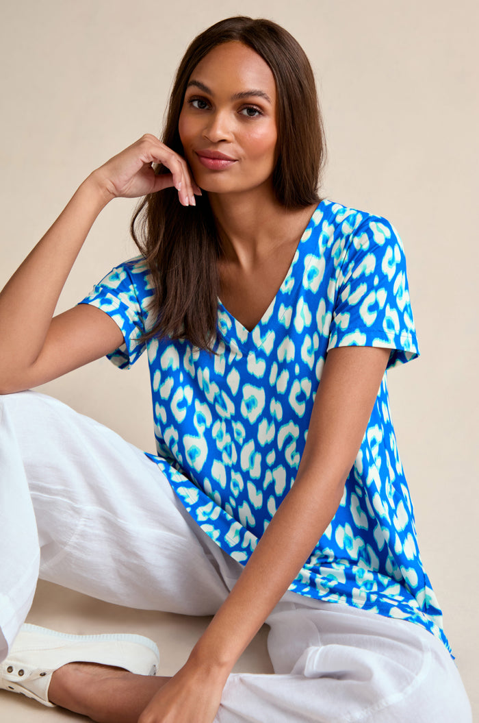 Woman seated against a beige background, wearing the Abstract Leopard Print Jersey Top in blue and white, paired with white pants and sneakers, with one hand resting on her face.