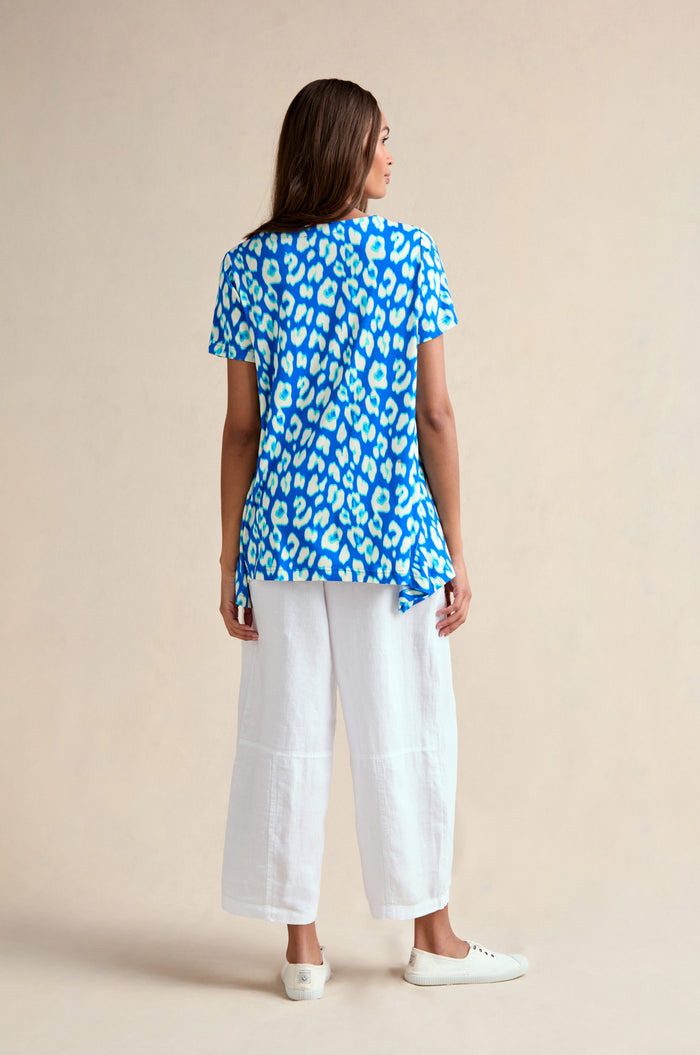 A woman stands facing away, wearing an Abstract Leopard Print Jersey Top with white wide-leg pants and white sneakers against a plain beige background.