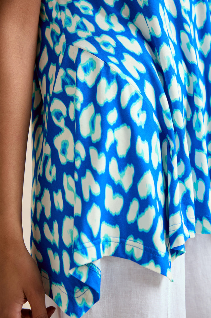 Close-up of a person wearing the Abstract Leopard Print Jersey Top, featuring a blue and white animal print and an asymmetrical hem, paired with white pants. Only part of the person's arm and hand are visible.