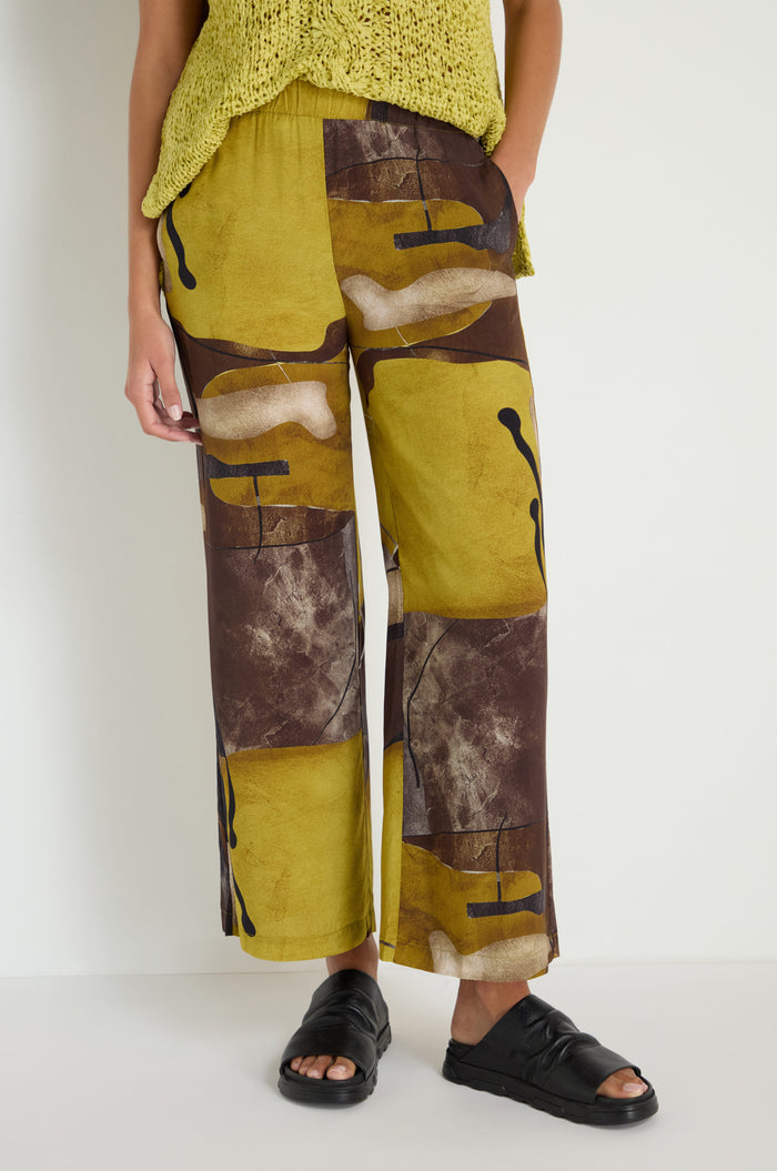 A person stands against a plain white wall wearing Abstract Print Trousers in yellow and brown, paired with a yellow knit top and black sandals.