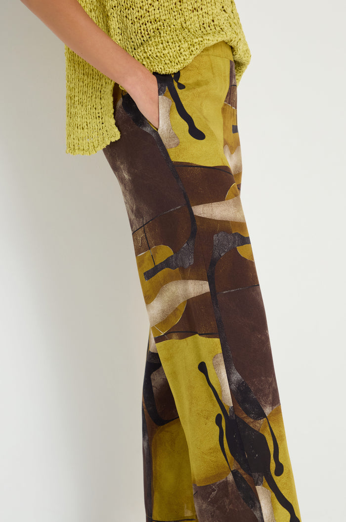 Person wears a yellow-green knit top and Abstract Print Trousers in brown, yellow, and black viscose, standing with one hand in pocket against a plain background.