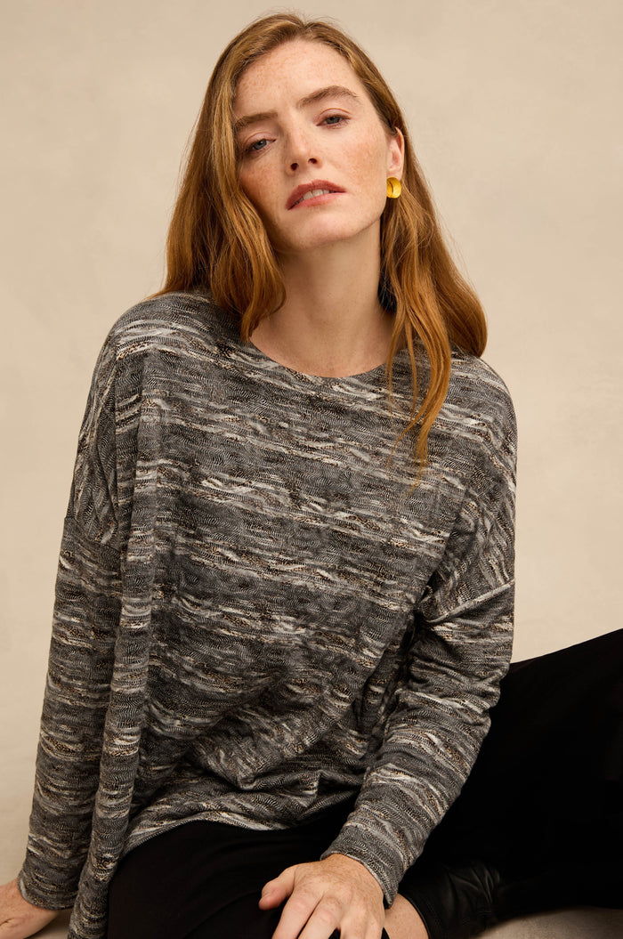 A woman with long red hair is wearing the Abstract Ripple Jersey Boxy Top—gray with a patterned design—paired with black pants and yellow earrings, seated against a light beige background.