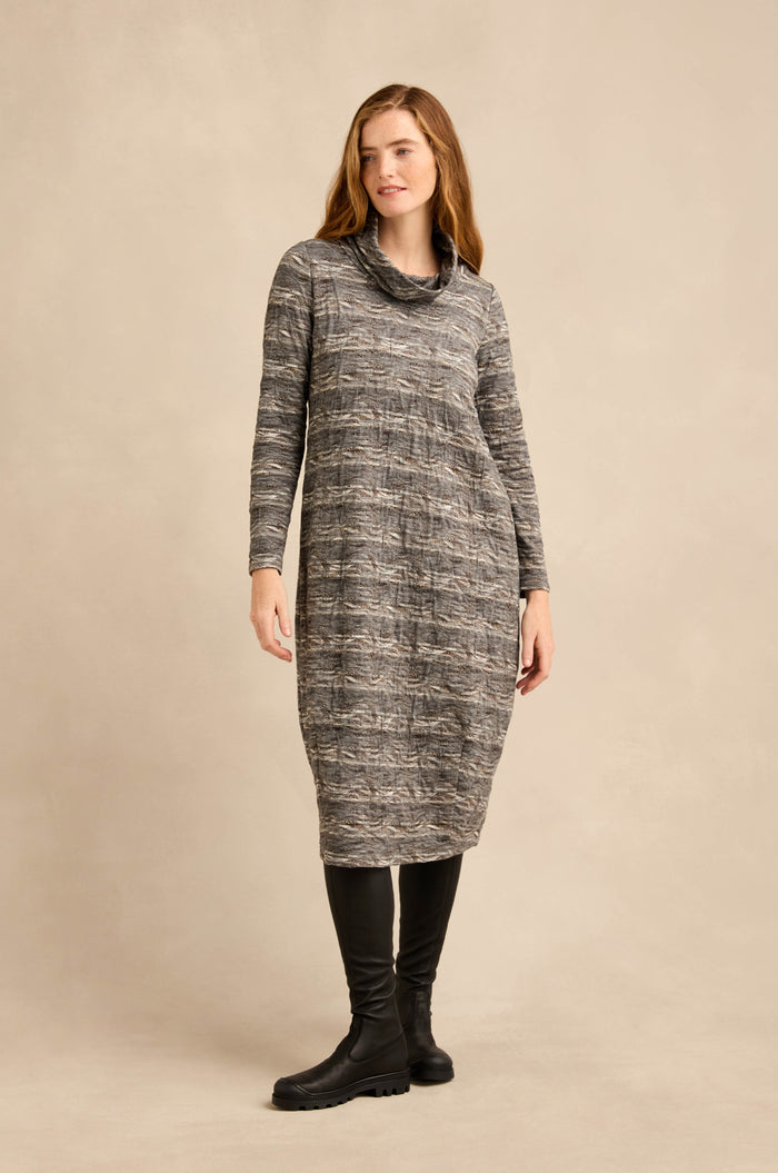 A woman stands against a plain beige background wearing the Abstract Ripple Jersey Bubble Dress in gray, featuring long sleeves, a subtle bubble silhouette, and paired with black knee-high boots.