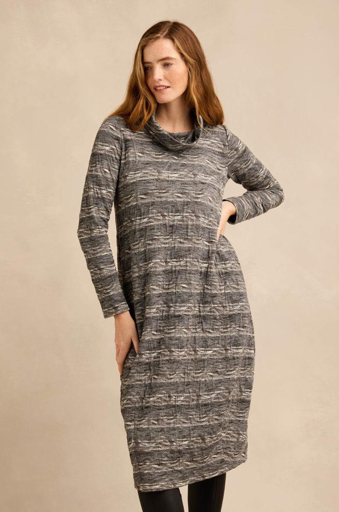 A woman stands with one hand on her hip against a neutral background, wearing the Abstract Ripple Jersey Bubble Dress—a long-sleeve, patterned gray textured dress featuring a cowl neck.