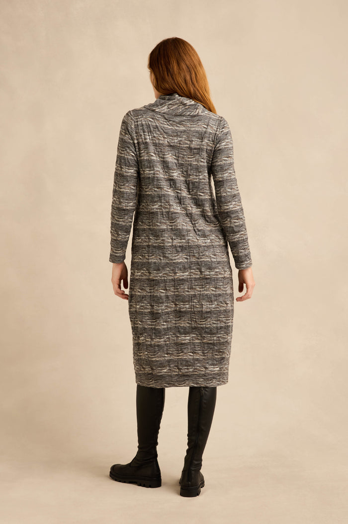 A person stands facing away, wearing a long-sleeved, knee-length gray Abstract Ripple Jersey Bubble Dress with a textured jersey finish and black leggings with black boots, against a beige background.