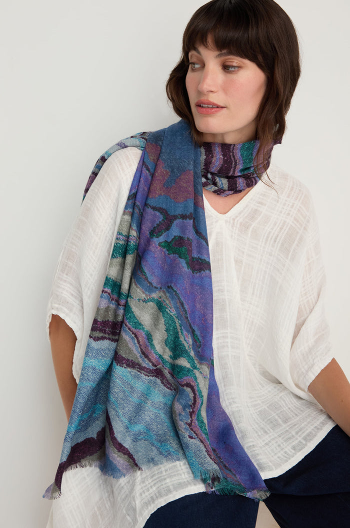 A woman seated against a neutral background wears a textured white top paired with the Agate Wool Scarf, which features an agate-inspired print in vibrant blue tones.