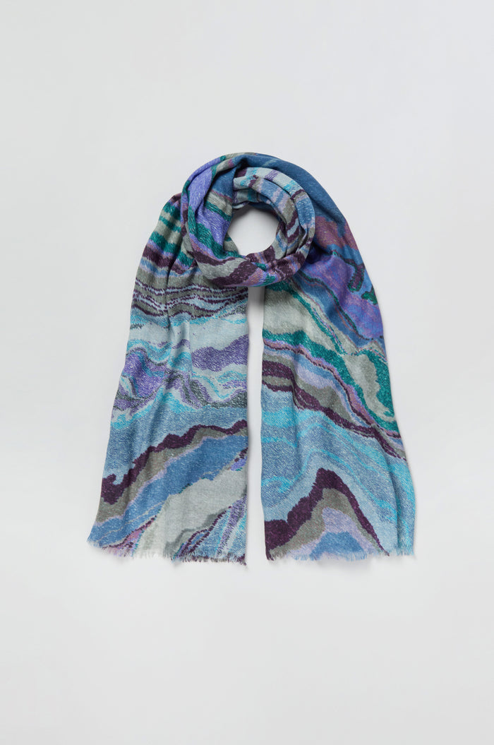 The Agate Wool Scarf is a luxurious wool-silk accessory featuring an agate-inspired marbled print in shades of blue, teal, purple, and gray, displayed flat against a light background.