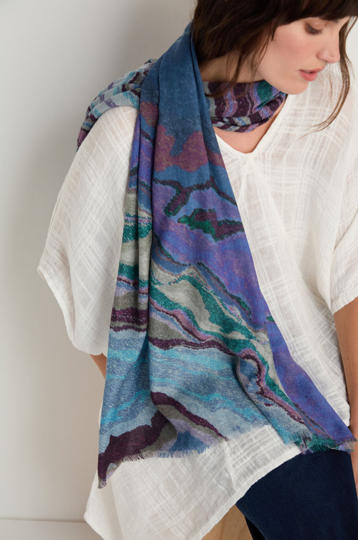A woman stands against a plain light background, wearing a loose white textured top paired with the Agate Wool Scarf—a blue wool-silk accessory featuring a green and purple agate-inspired print.