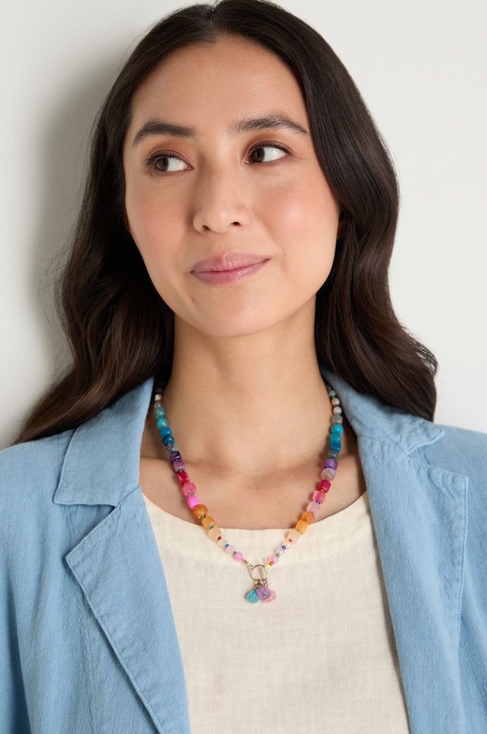 A woman with long dark hair wears a light blue blazer, cream top, and the Artisanal Rainbow Agate Toggle Necklace, handmade in Scotland, while looking to the side against a plain background.