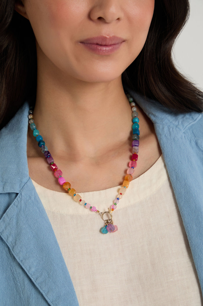 A woman wearing a light blue blazer and cream top showcases the Artisanal Rainbow Agate Toggle Necklace, handmade in Scotland with natural agate stones and charms.