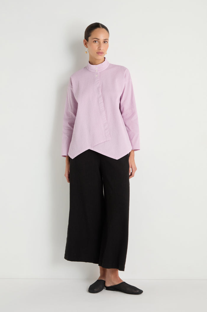 A woman stands against a white wall wearing the Asymmetric Cotton Lilac Shirt, featuring stitched stripes, paired with wide-leg black pants and black flat shoes.