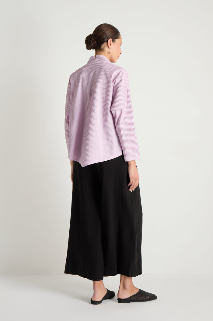 A person stands facing away, wearing an Asymmetric Cotton Lilac Shirt, loose black pants, and black slip-on shoes against a plain white background.