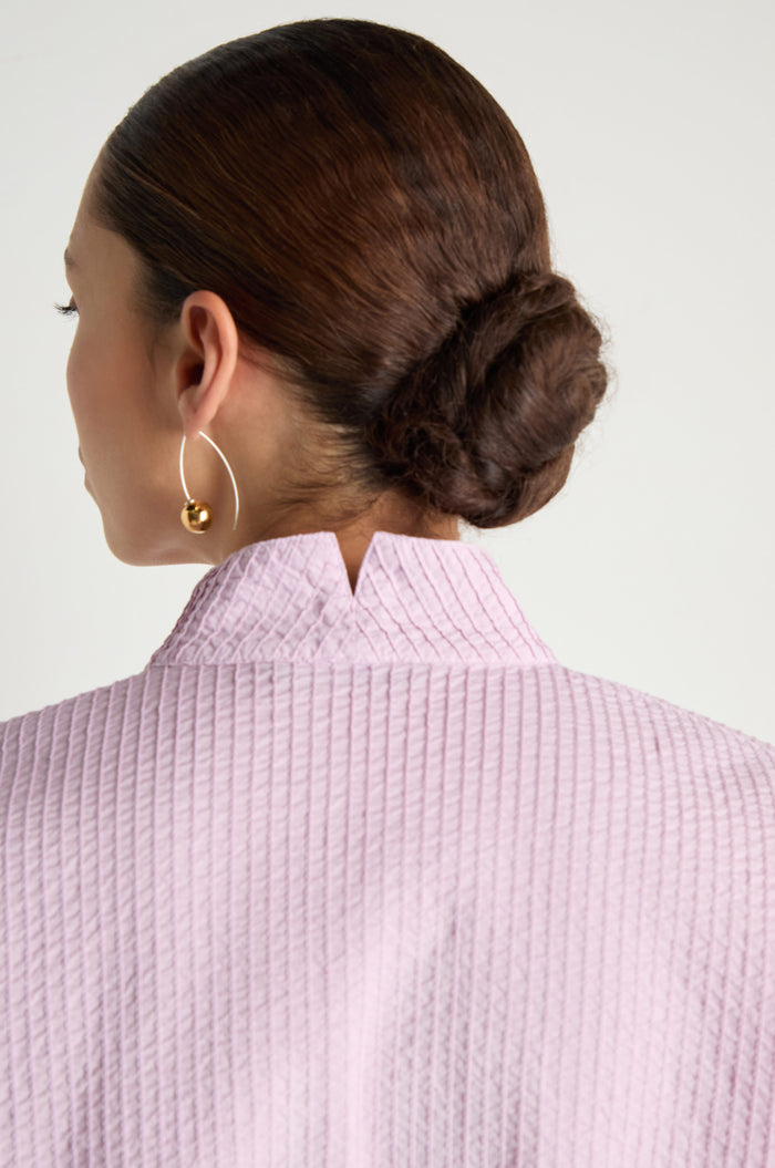 A person with brown hair in a low bun, shown from the back, wears an Asymmetric Cotton Lilac Shirt with a notched collar and stitched stripes, paired with a gold hoop earring.