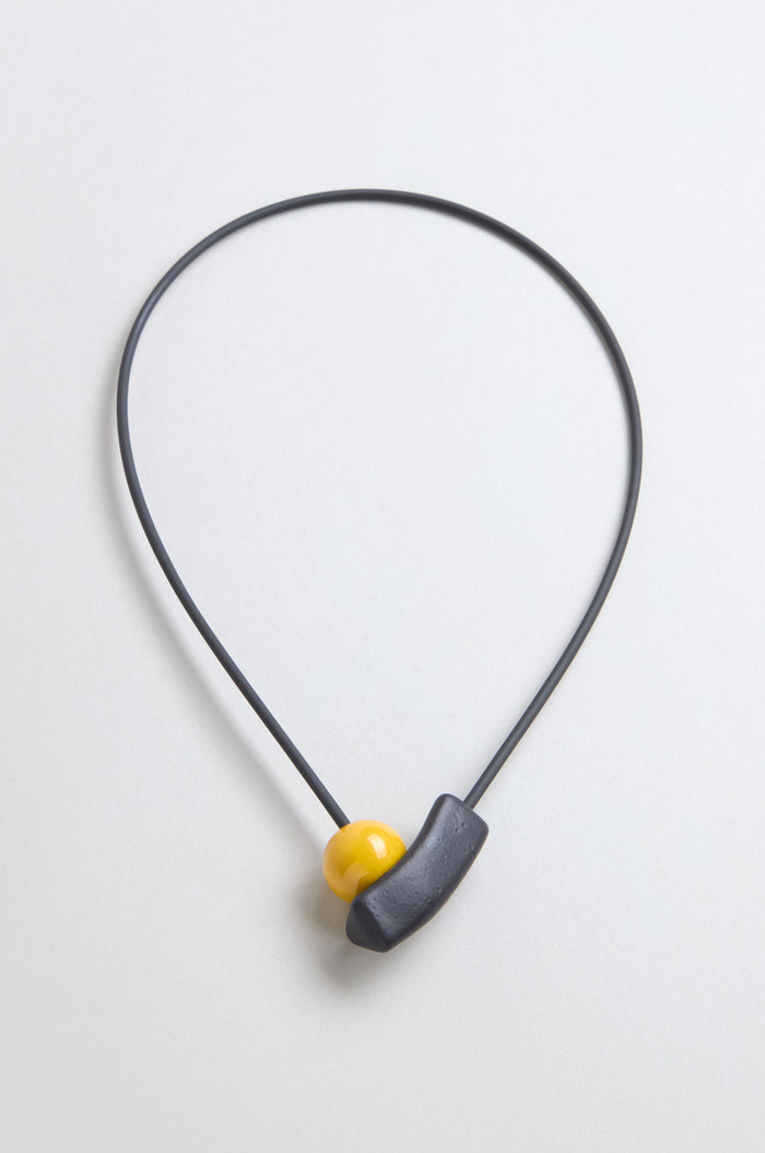 The Atom Necklace is a minimalist ceramic accessory with a thin black cord, featuring geometric elements—a yellow bead and curved black piece—displayed against a light background.