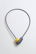 The Atom Necklace is a minimalist ceramic accessory with a thin black cord, featuring geometric elements—a yellow bead and curved black piece—displayed against a light background.