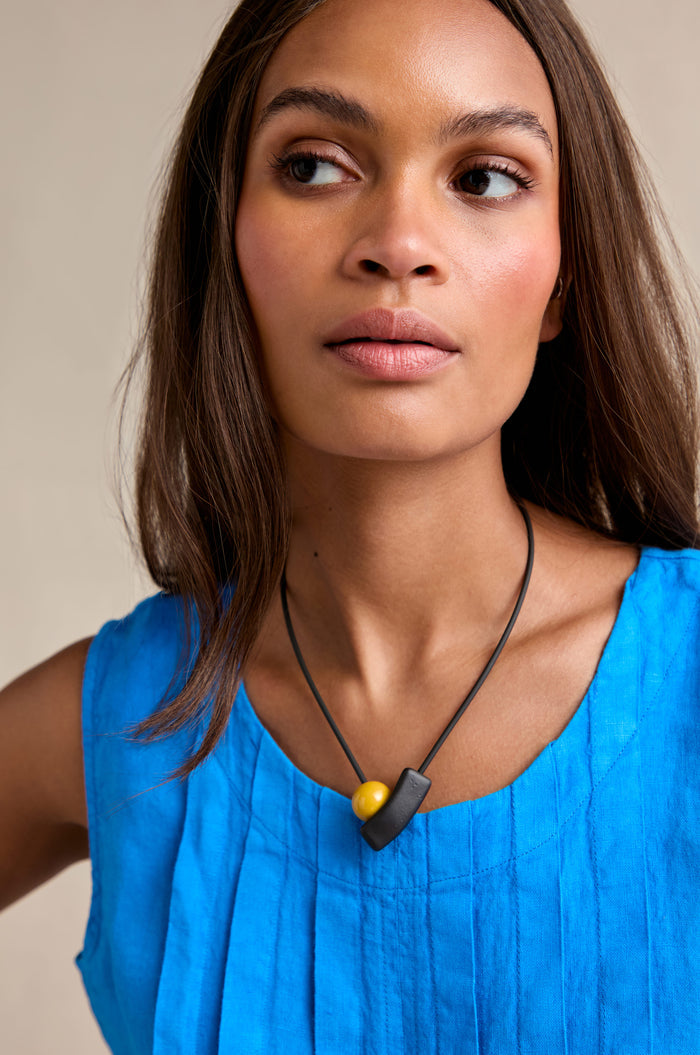 A woman in a sleeveless blue top wears the Atom Necklace, featuring geometric black and yellow enamel beads, as she looks to the side against a neutral background.
