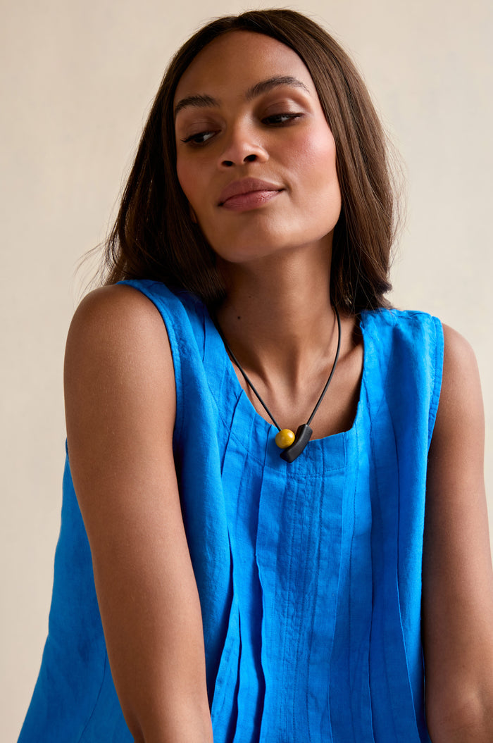 A woman with straight brown hair wears a sleeveless bright blue top and the Atom Necklace—featuring black geometric elements with yellow and black beads—looking down and to the side against a neutral background.