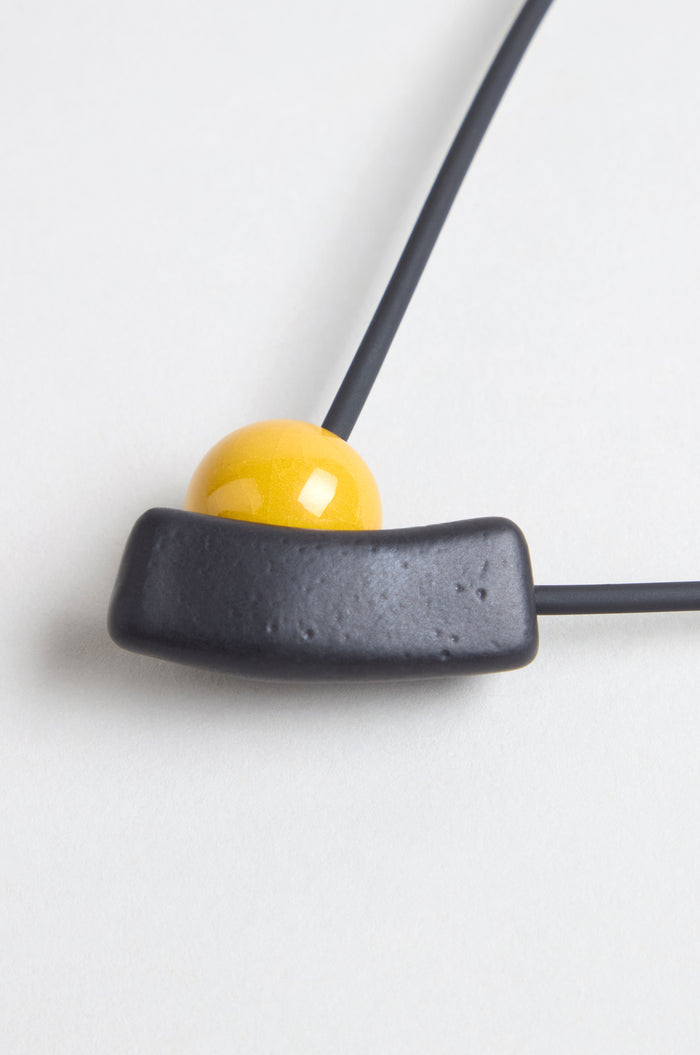 Close-up of the Atom Necklace, a ceramic piece with geometric design, featuring a yellow enamel spherical bead and a black curved rectangular pendant on a black cord, shown against a white background.