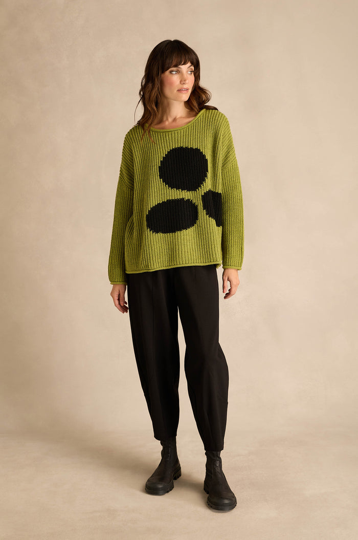 A woman stands against a neutral background, wearing the Avocado Spot Scoop Neck Knit with black pants and black boots.