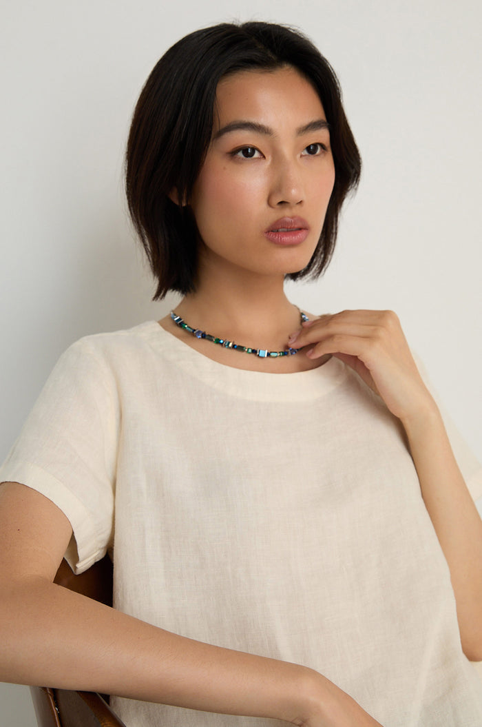 A person with short dark hair, wearing a light-colored top and the Blue Hematite Necklace, sits against a plain background looking slightly to the side.