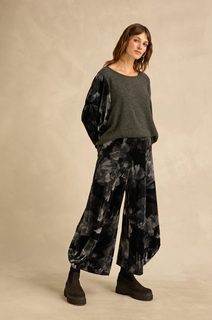 Woman standing against a beige background, wearing a loose grey sweater with sculptural volume, Blurred Floral Velvet Bubble Trousers, and black platform boots.