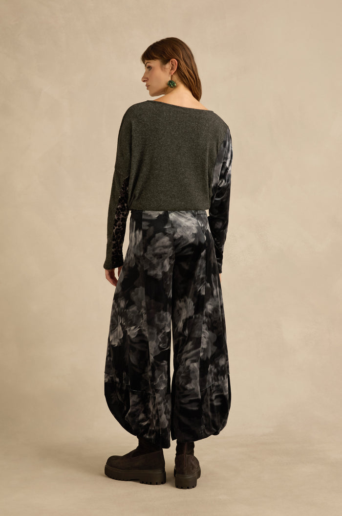 A person stands with their back to the camera, wearing a dark gray long-sleeve top, Blurred Floral Velvet Bubble Trousers in black-and-gray tie-dye with sculptural volume, platform boots, and green earrings against a beige background.