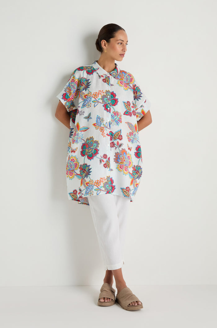 A woman stands against a white wall wearing the Botanical Linen Long Shirt, paired with white pants and beige slides.