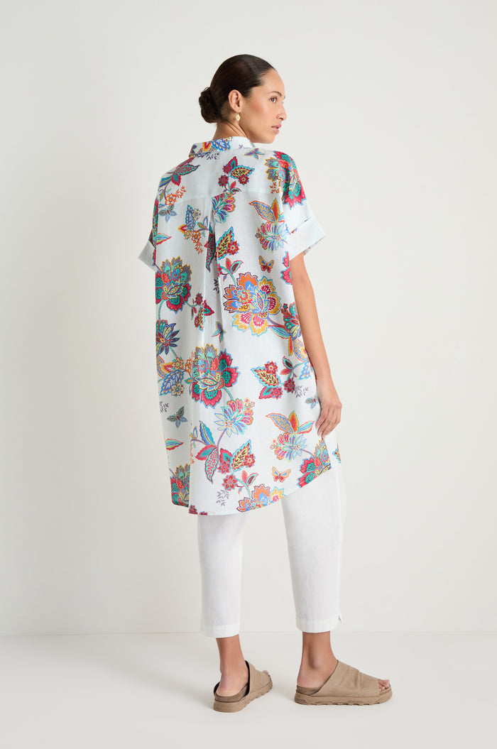 A woman stands sideways against a plain white background, wearing the Botanical Linen Long Shirt with white pants and beige sandals.