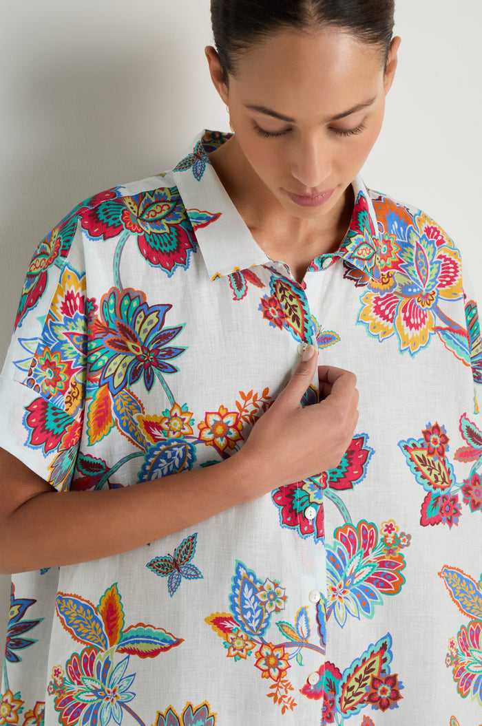 A woman wearing the Botanical Linen Long Shirt in a white botanical print holds the collar with her right hand while looking down.