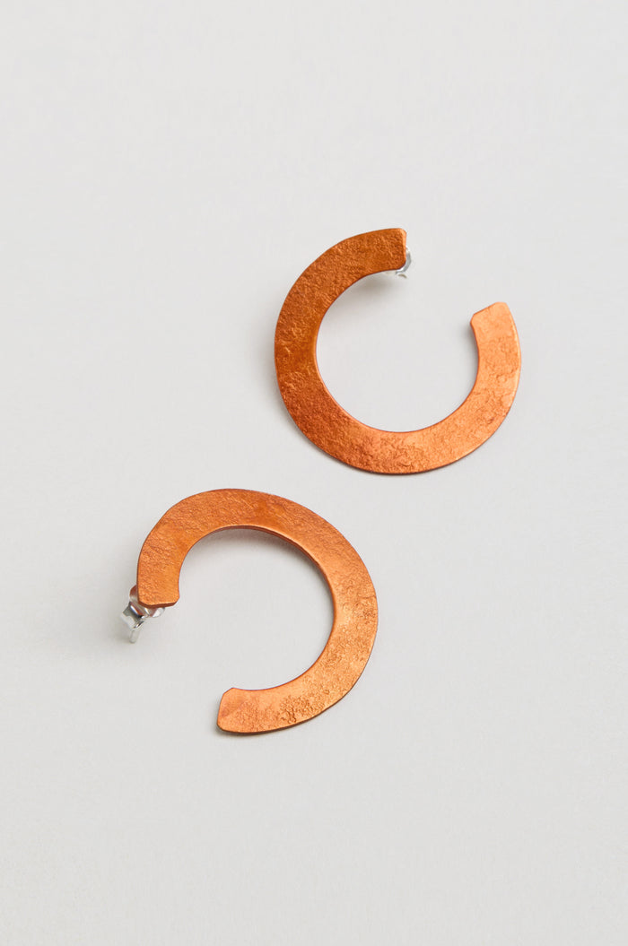 The C Curve Earrings feature a modern hoop shape in a distinctive C design, crafted from textured copper-colored metal and shown against a plain light background.
