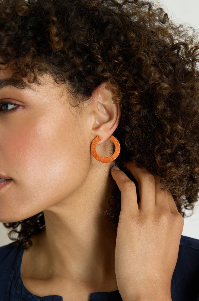 A person with curly hair touches a C Curve Earring on their left ear, with part of their face and hand visible against a neutral background.