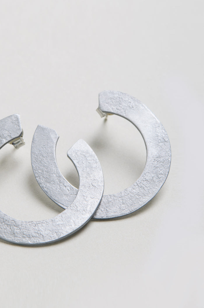 The C Curve Earrings feature a textured, silver-toned finish and a modern hoop design, displayed on a light-colored surface.