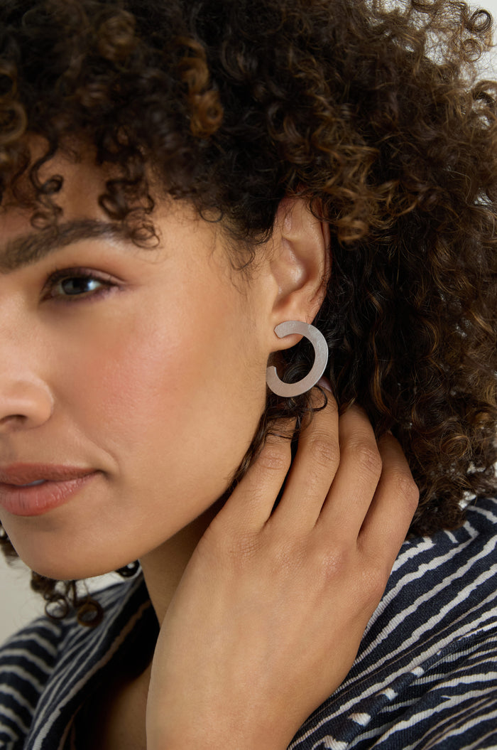 A woman with curly hair and a striped top touches her ear, showcasing C Curve Earrings in this close-up portrait.