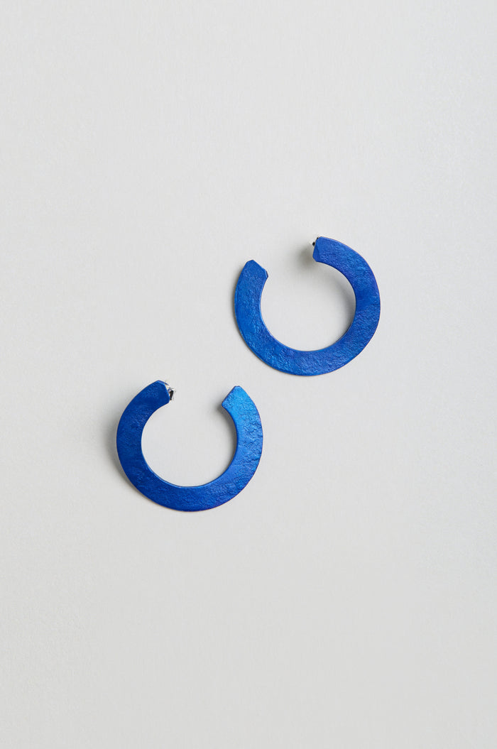 A pair of C Curve Earrings—round, flat, open-ended hoops in blue anodised aluminium—are displayed on a light grey surface.