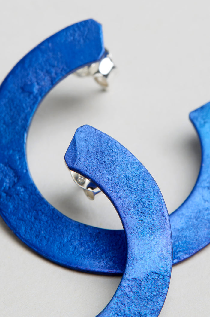 Close-up of two textured C Curve Earrings in blue anodised aluminium with silver posts, displayed on a light gray background.