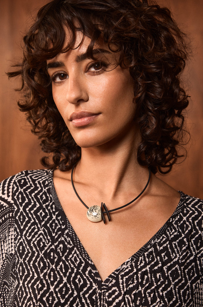 A woman with short curly brown hair wears a black and white patterned top and the handmade Calligraphy Necklace featuring a round ceramic pendant, standing in front of a wooden background.