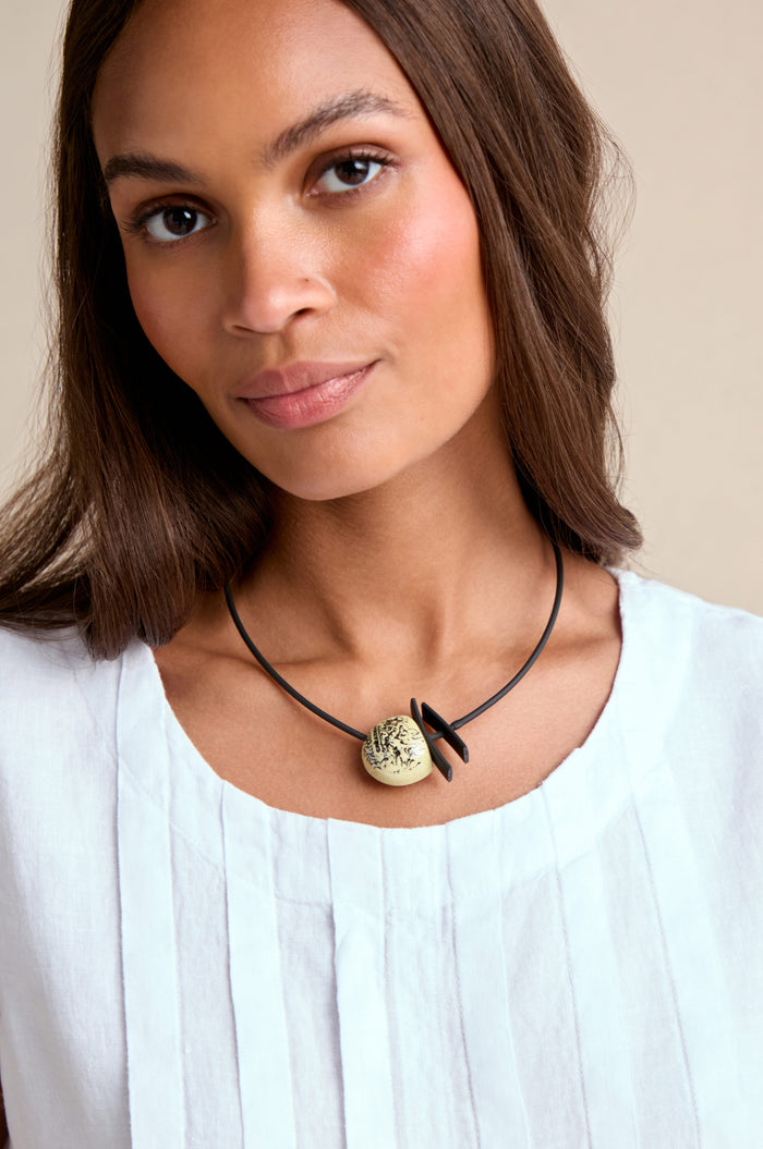 Woman with straight brown hair wears a white pleated top and the Calligraphy Necklace, a black cord featuring a round beige ceramic pendant.