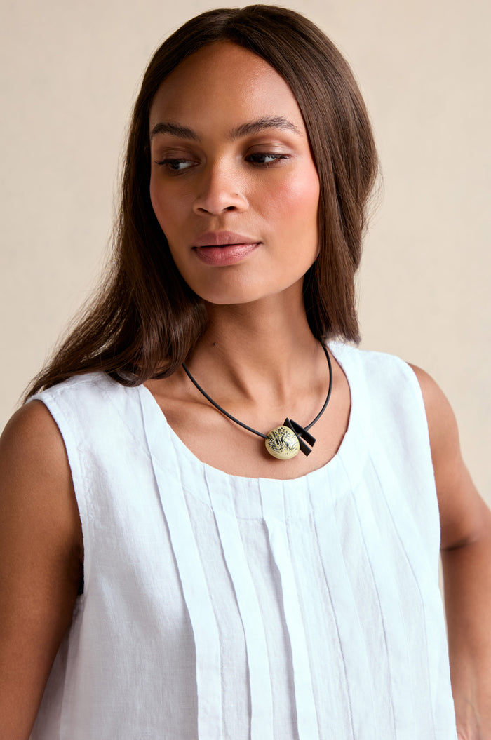 Woman with straight brown hair wears a sleeveless white pleated top and the Calligraphy Necklace—a ceramic pendant on a black cord—while looking slightly to the side against a neutral background.