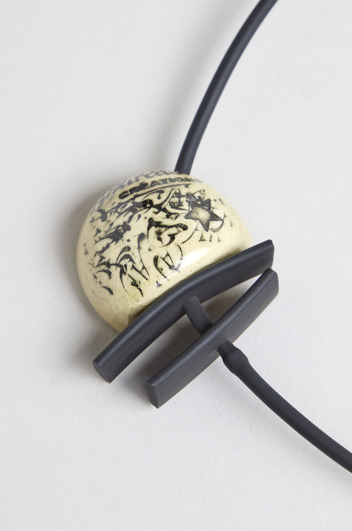 The Calligraphy Necklace features a round cream ceramic pendant with black abstract markings, accented by two matte black curved bars on a black cord.
