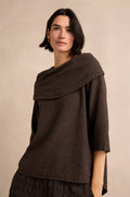 A woman with short dark hair wears the Chocolate Draped Neck Top—crafted from textured brown cotton-viscose jersey and featuring a wide, asymmetric cowl collar—as she stands against a neutral beige background.