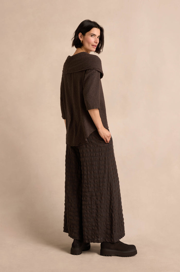 A woman stands sideways against a neutral background, wearing the Chocolate Draped Neck Top—crafted from a loose brown cotton-viscose jersey with wide sleeves and an elegant, asymmetric cowl neckline for added sophistication—paired with matching wide-leg pants and black platform shoes.