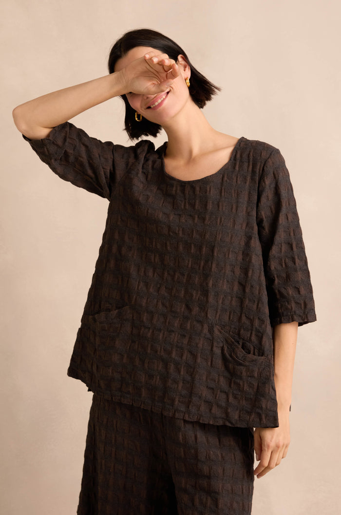 Wearing the Chocolate Waffle Check Flared Top with matching pants, a person stands against a beige background, smiling and raising one arm to partially cover their face.