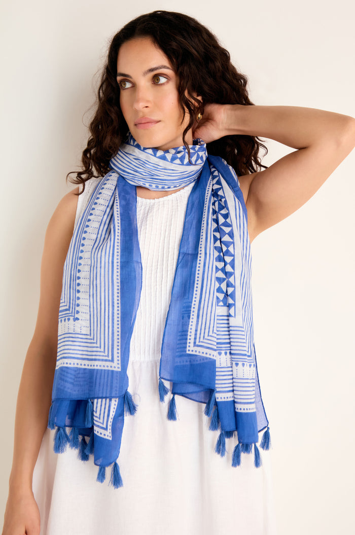 A woman stands against a plain light background, wearing the Cobalt Tassel Cotton Scarf featuring a blue and white geometric print and tassel edging, draped over a sleeveless white dress.