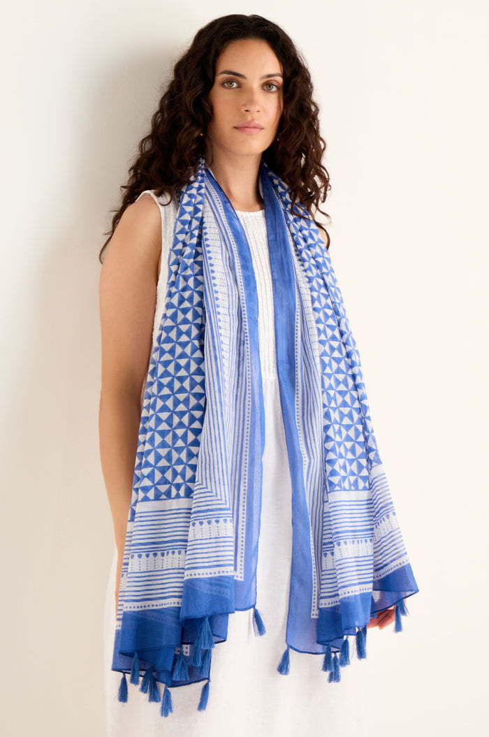 A woman with long curly hair, dressed in a white dress, models the Cobalt Tassel Cotton Scarf—a blue geometric print cotton scarf finished with tassel edging—against a plain light background.