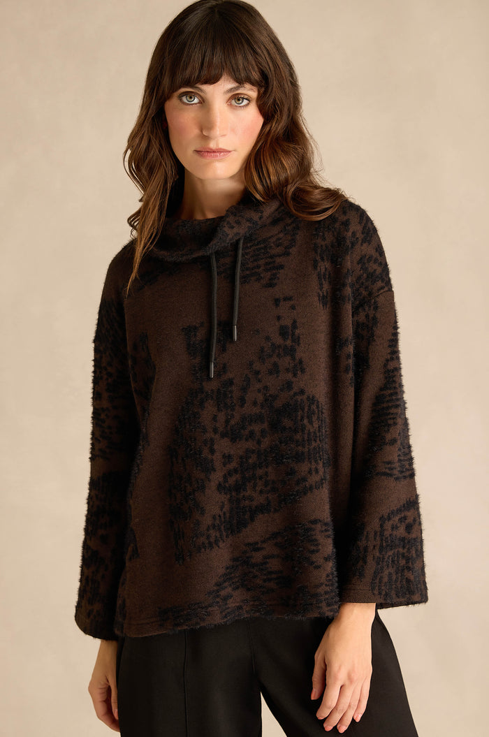 A woman wearing the Cocoa Speckled Jersey Boxy Top—a loose, long-sleeve, dark brown boxy fit top with a cowl neck, black abstract pattern, and black drawstrings—paired with black pants.